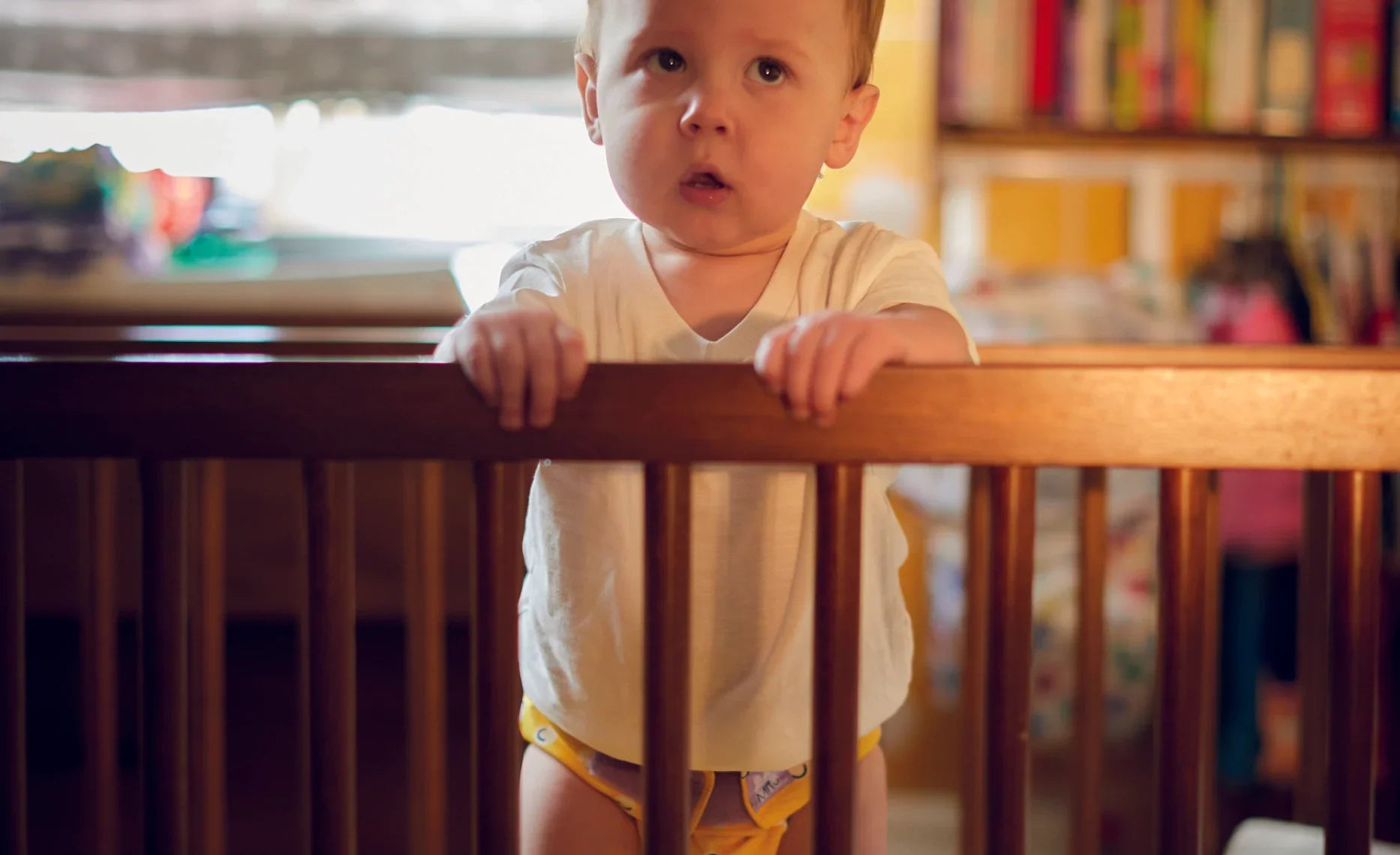 Baby standing in a crib with a blurred background of books and toys